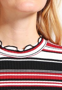 Kobieta z blond włosami, widok na szyję i dolną część twarzy, ubrana w żebrowany, paskowany top w kolorach czerwonym, białym i czarnym z falbankami na dekolcie.