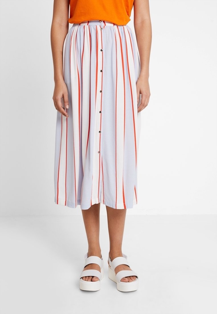 Bílá midi sukně s červenými vertikálními pruhy, zapínáním na knoflíky vpředu a elastickým pasem, doplněná oranžovým topem a bílými sandály na platformě.