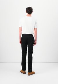 Sorte bukser med slim fit, lavet af bomuldsstof, to baglommer, båret med en hvid t-shirt og brune sko, set fra ryggen.