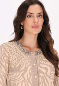 Beżowy kardigan dziergany z jasnobrązowym wzorem zebry, ozdobiony złotymi i srebrnymi ćwiekami, z biżuteryjnym kołnierzem i zapięciem na guziki.