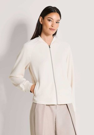 Weiße Reißverschlussjacke mit Raglanärmeln, ausgestattet mit Seitentaschen und einer glatten, weichen Textur. Kombiniert mit hellbeigen, weit geschnittenen Hosen.