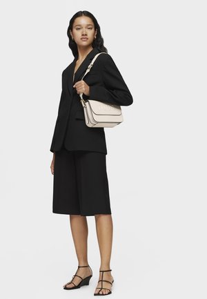 Bolso de cuero beige con el logo "TOUS" en relieve, con ribetes negros, una correa para el hombro y forma rectangular, combinado con un traje negro y sandalias de tacón.