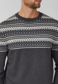 Homme portant un pull gris charbon avec un empiècement à motifs comportant des designs géométriques et floraux en blanc et gris clair.