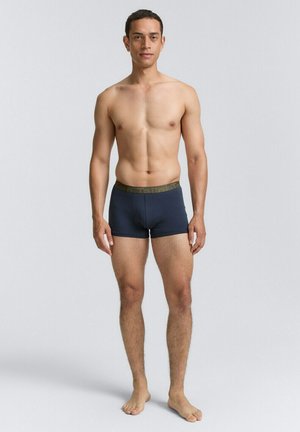 Herren Boxershorts in Marineblau mit einem strukturierten grünen Bund, figurnaher Schnitt und weichem Stoff, getragen von einem Model, das barfuß steht.