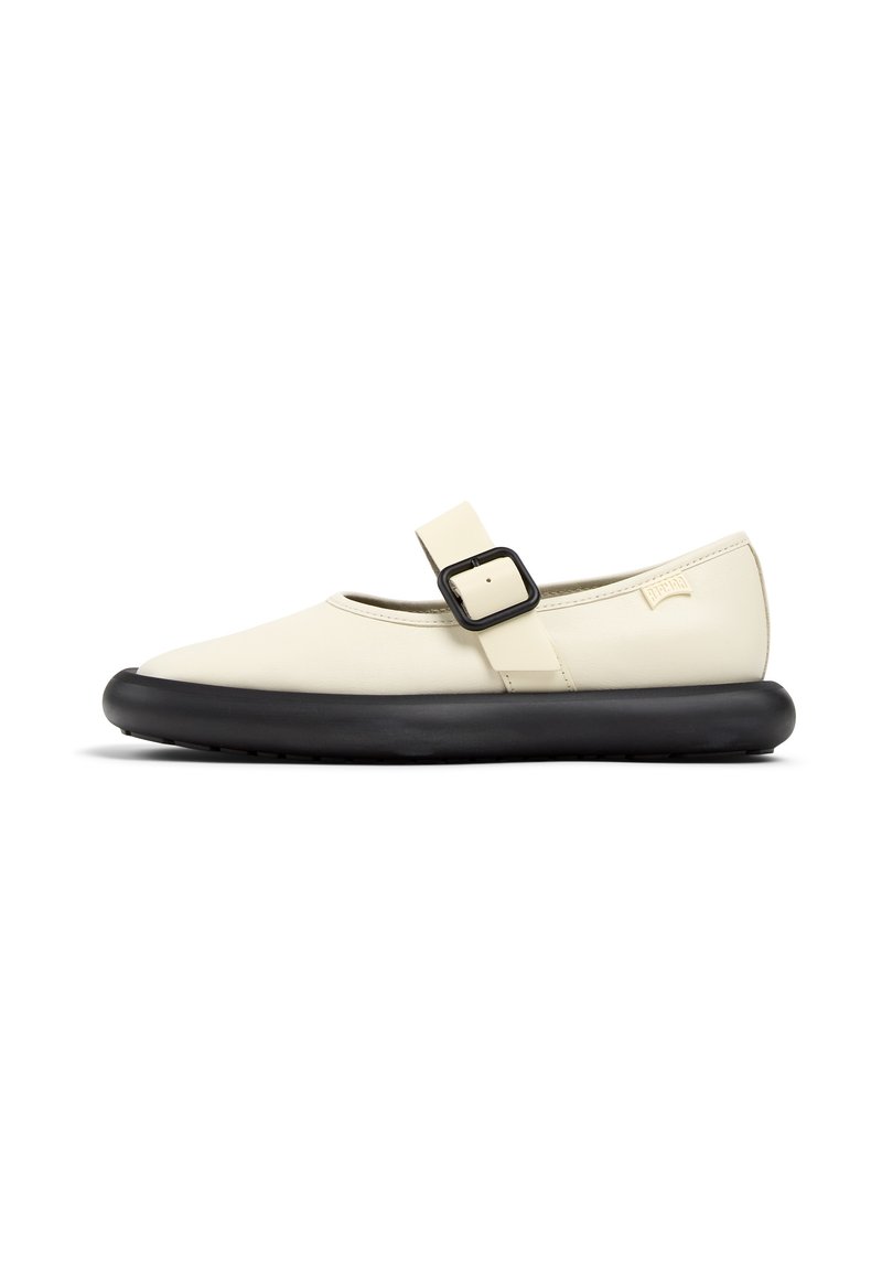Zapato ligero de color crema con punta redondeada, suela de goma negra y una hebilla de correa negra cruzando el pie. Textura suave.