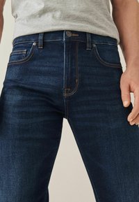 Donkerblauwe denim jeans met een slanke pasvorm, voorzien van oranje stiksels, een traditionele vijfzakkenontwerp en een enkele knoopsluiting.