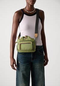 Zaļa neilona crossbody soma ar divām augšējām rokturiem, regulējamu siksniņu, priekšējo rāvējslēdzēja kabatu un zelta aparatūras akcentiem. Strukturēta virsma, kompakta forma.