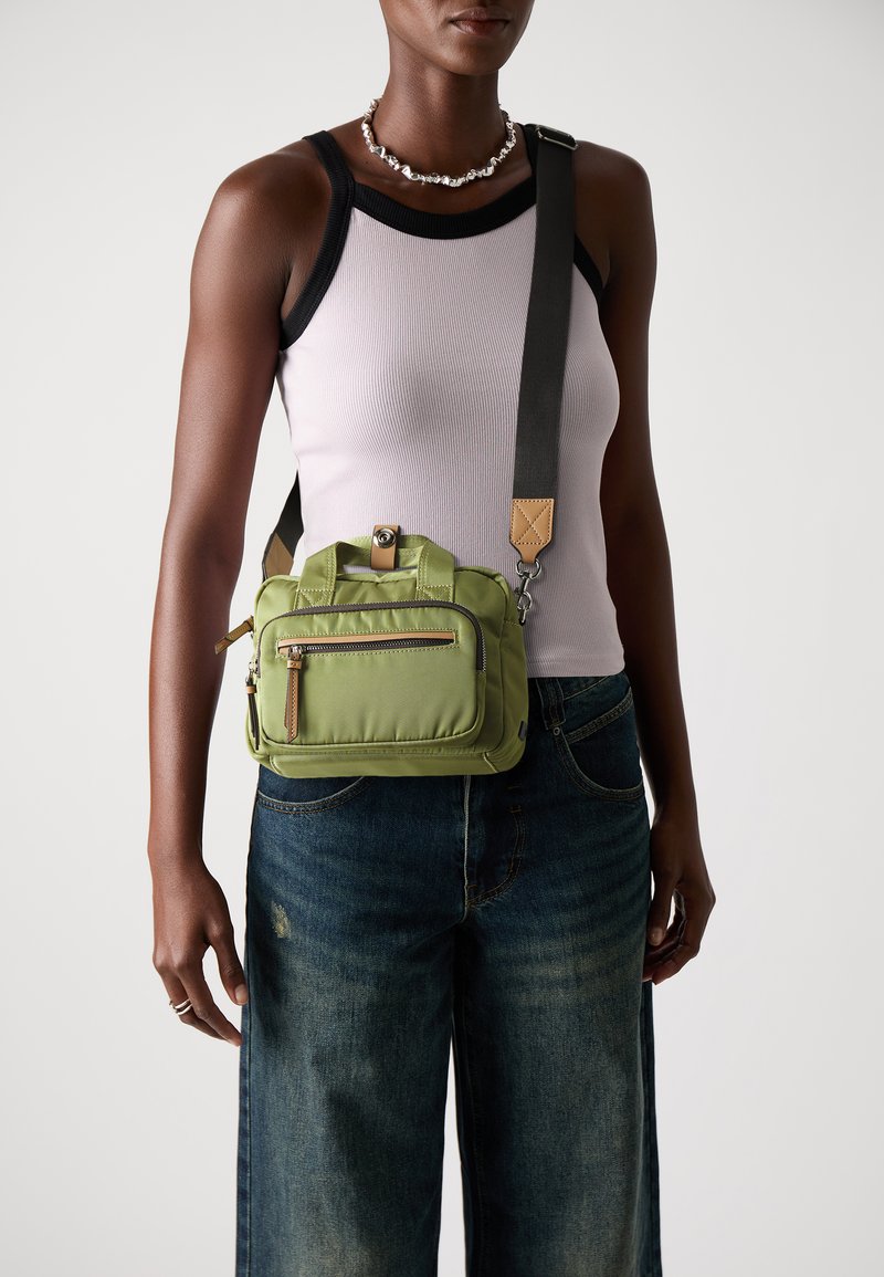 Zaļa neilona crossbody soma ar divām augšējām rokturiem, regulējamu siksniņu, priekšējo rāvējslēdzēja kabatu un zelta aparatūras akcentiem. Strukturēta virsma, kompakta forma.