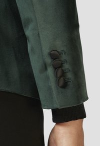 Ärmel einer grünen Samtjacke mit strukturiertem Finish, versehen mit vier runden schwarzen Knöpfen und einem schwarzen gerippten Bündchen.