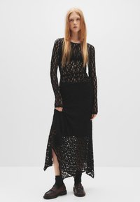 Top in pizzo nero con maniche lunghe e collo alto abbinato a una gonna nera asimmetrica al ginocchio, con fondo trasparente e tessuto texturizzato.