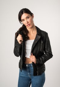 Schwarze Lederbikerjacke mit Reißverschlusstaschen und Schulterklappen. Getragen über einem weißen gerippten Crop-Top und kombiniert mit blauen High-Waist-Jeans.