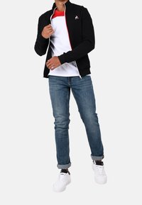 Chaqueta negra con cremallera sobre una camisa de capas blanca y roja, combinada con vaqueros azules y zapatillas blancas. Presenta un logotipo en la chaqueta.