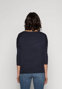 Mörkblå stickad tröja med trekvartsärmar, ribbade muddarna och rund halsringning, tillsammans med tajta blå jeans. Detaljerad sömnad synlig.