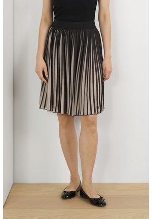 Femme portant une jupe noire plissée mi-longue avec doublure beige, un haut noir et des ballerines noires, debout sur un sol en bois.