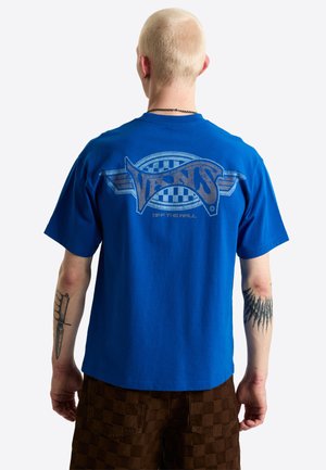 Persona con cabello corto y rubio que lleva una camiseta azul de Vans "Off The Wall" y pantalones cortos marrones a cuadros, vista desde atrás.