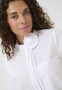 Camicia bianca con bottoni, con un riflesso floreale arricciato al colletto, dotata di una tasca sul petto e una superficie in tessuto liscia.