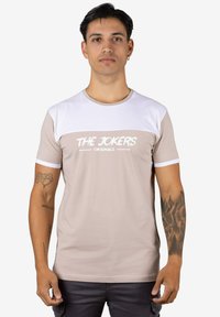 Camiseta de manga corta en beige y blanco, con cuello redondo, textura suave y texto en negrita "THE JOKERS ORIGINALS" en el frente.