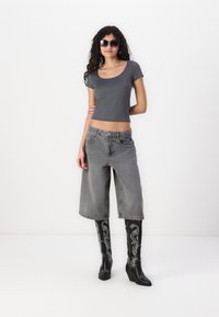 T-shirt gris coupé, culottes en denim gris amples, bottes compensées noires brodées jusqu'aux genoux et lunettes de soleil noires. Tissu lisse et textures contrastantes.