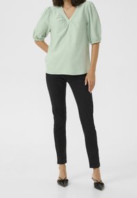 Blouse à manches courtes vert clair avec col en V et tissu texturé ; associée à un pantalon noir slim et des ballerines noires pointues.