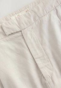 Pantalons gris clair présentant une texture lisse, une taille plate et des détails cousus, avec une étiquette visible à l'intérieur de la ceinture.