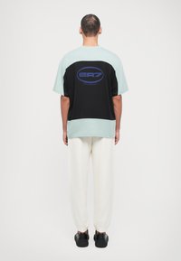 Μαύρο και μέint πράσινο κοντομάνικο T-shirt με λογότυπο στην πλάτη, συνδυασμένο με άσπρα παντελόνια jogging και μαύρα αθλητικά παπούτσια, απεικονιζόμενο από πίσω.