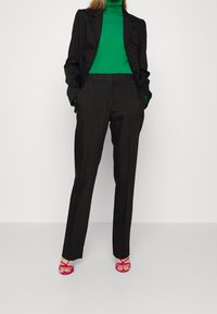 Svart skräddarsydd kavaj över en grön polotröja, matchad med högmidjade svarta byxor och rosa sandaletter med klack. Släta texturer och skarpa linjer.