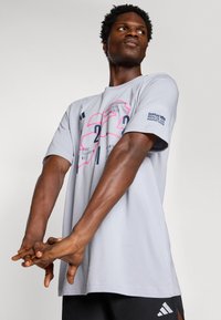 T-shirt de sport gris avec des manches courtes, présentant des impressions graphiques en rose et noir, ainsi que le logo du Marathon de Berlin BMW sur la manche.