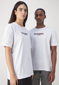 Camisetas de algodón blancas con el logo de "Tommy Jeans" en negro y rojo, con un corte relajado y mangas cortas. Diseño sencillo y textura suave.