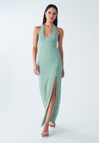 Robe longue vert menthe texturée avec un col licou et une fente latérale. Présente un corsage ajusté et une jupe fluide, complétée par des sandales blanches.