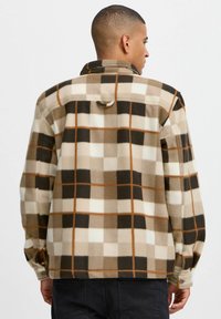 Fleece overskjorte i brun, beige og hvit rutete mønster. Har knapper, krage og løkke detalj på baksiden. Myk tekstur.