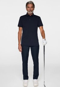 Granatowa koszulka polo z kołnierzykiem, zestawiona z ciemnymi spodniami. Model trzyma kij golfowy, ma na sobie białą rękawiczkę i jasne buty.