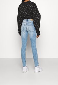 Sweat-shirt noir à motifs circulaires dorés, accompagné de jeans skinny bleu clair. Le mannequin porte des baskets blanches. Texture lisse, coupe ajustée.