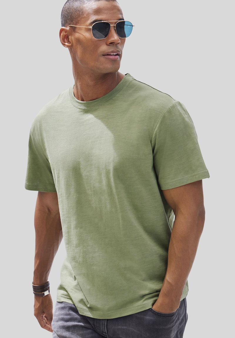 Olive green short-sleeve t-shirt in soft fabric with a round neckline, paired with dark grey pants and a brown wristband.