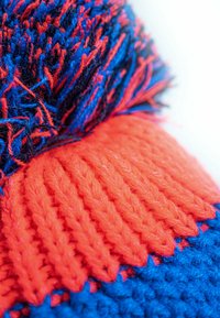 Knit hat with vibrant blue and orange yarn, featuring a textured ribbed band. A fluffy pom-pom sits atop, showcasing varied yarn lengths.