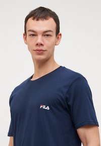 Tume sinine puuvillane T-särk, millel on ümar kaelus, lühikesed varrukad ja vasakul rinnal valge FILA logo punaste aktsentidega.