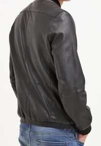 Giacca bomber in pelle nera perforata con orlo e polsini a costine, caratterizzata da una finitura liscia e cuciture dettagliate sul retro.