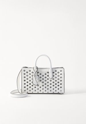 Sac à main en cuir blanc avec œillets métalliques noirs, deux poignées, une bandoulière et une étiquette suspendue sur un fond blanc uni.