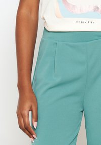 Pantalones color verde azulado con plisados, hechos de una tela suave y lisa. Detalles visibles en la cintura y la costura lateral. Una mano que descansa cerca muestra un diseño de uñas neutro.