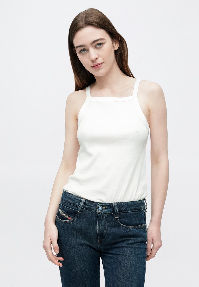 Young woman with long brown hair wearing a white sleeveless top and dark blue jeans, standing against a plain white background.