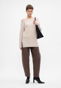 Beige oversized pletený sveter s dlhými rukávmi, v kombinácii s hnedými voľnými nohavicami a čiernymi čižmami na vysokom opätku. Čierna kabelka v ruke.