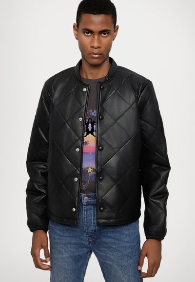 Veste en cuir matelassé noire avec un col rond, des boutons-pression et des poignets élastiques, portée sur un t-shirt graphique et un jean bleu.