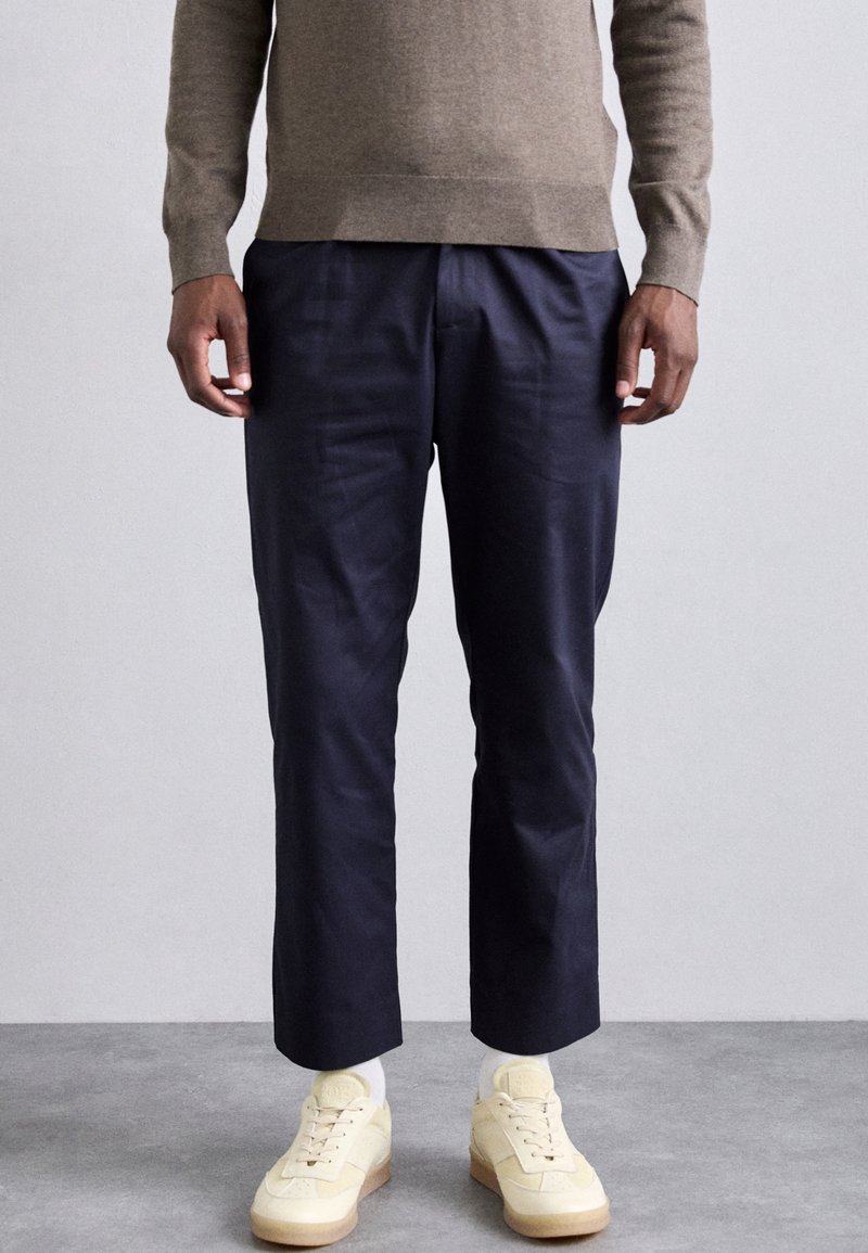 Marineblauwe broek van gladde stof met een getailleerde pasvorm, gecombineerd met crèmekleurige sneakers met een textuurdesign en lichte tinten.