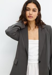 Grauer Blazer mit strukturierten Revers, Ein-Knopf-Verschluss und seitlichen Taschen, kombiniert mit einem weißen trägerlosen Oberteil. Glatter Stofftextur.