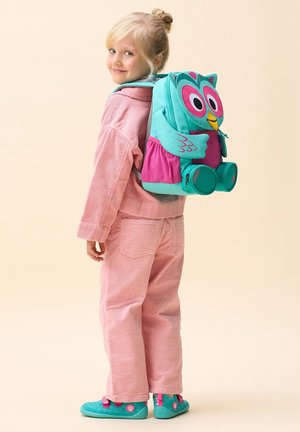 Junges Mädchen in pinkem Outfit mit türkis-pinkfarbenem Eulenkinder-Rucksack, lächelnd und über die Schulter blickend.