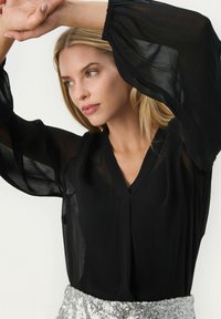 Blusa nera trasparente con scollatura a V e maniche voluminose, caratterizzata da una silhouette fluida. Abbinata a una gonna di paillettes argento.