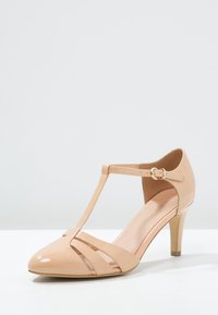 Béžová lakovaná T-pásková vysoká podpatková bota s špičatou špičkou, dekoračními výřezy a spona na kotníkovém pásku.