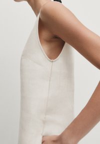 Seitenansicht einer Person, die ein ärmelloses, cremefarbenes, strukturiertes Kleid mit runder Armausschnitt vor einem schlichten Hintergrund trägt.