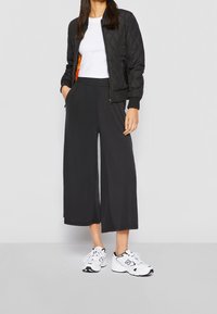 Frau trägt eine schwarze gesteppte Bomberjacke über einem weißen, bauchfreien Top, schwarze, weite, gekürzte Hosen und weiße Turnschuhe, steht mit den Händen in den Taschen.