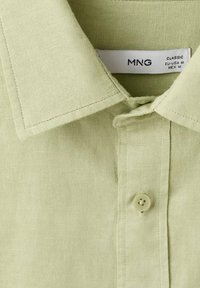 Camisa verde claro con cuello clásico, cierre de botones y tejido texturizado. La etiqueta muestra el nombre de la marca MNG y la información de la talla.