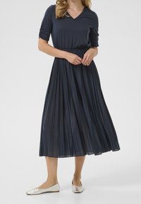 Robe plissée marine avec un col en V et des manches trois-quarts, accompagnée de ballerines argentées. Texture lisse et silhouette fluide.